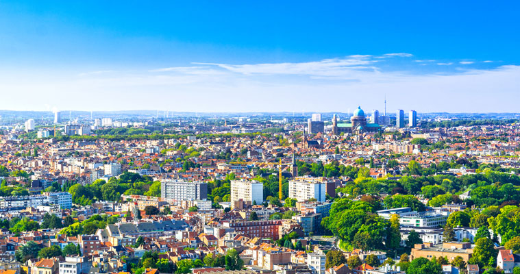 Panoramic cityscape featuring residential and commercial buildings, high-rises, a dome-shaped structure, and green trees under a bright blue sky with scattered clouds.