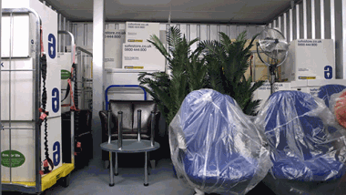 Opslagunit gevuld met gelabelde Safestore-dozen, stoelen, een tafel, een metalen kar, kunstplanten en een ventilator aan de muur.