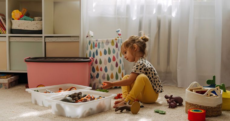 Enfant assis par terre jouant avec des jouets, entouré de boîtes de rangement et d’objets colorés dans une pièce lumineuse avec des rideaux clairs et des meubles.