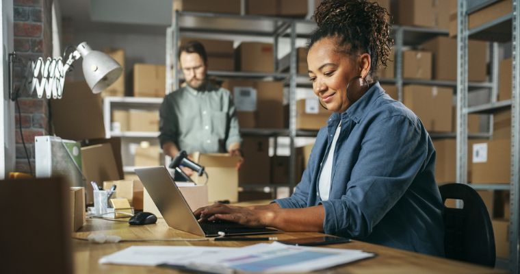 Deux personnes travaillant dans un entrepôt ou une zone de stockage avec des étagères et des cartons ; l’une est assise à un bureau avec un ordinateur portable, l’autre manipule un carton en arrière-plan.