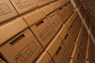 Storage room with rows of labeled cardboard archive boxes stacked on metal shelves, used for organized business stock or document storage.