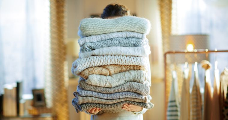 Personne tenant une grande pile de pulls pliés dans des tons neutres, le visage caché ; en arrière-plan flou, une lampe et des vêtements suspendus.