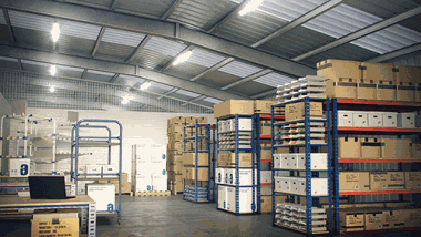 Warehouse interior with metal shelving units stacked with cardboard boxes, a blue trolley, and a workstation featuring a laptop.