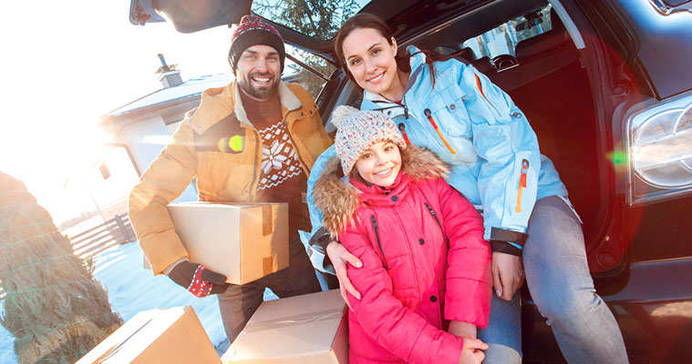 Drie personen in winterkleding bij een open kofferbak, die kartonnen dozen inladen in een besneeuwd landschap met bomen en een huis op de achtergrond.