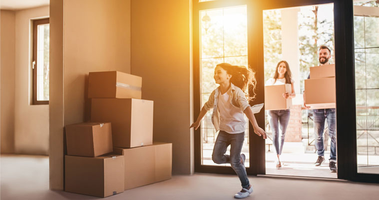 Child running inside a room with stacked cardboard boxes, while two adults enter through a glass door carrying more boxes, suggesting a home move-in scene.