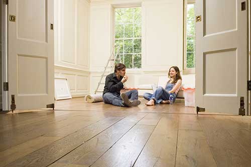 Persoon zit op de vloer in een woonkamer en sorteert kleding in een kartonnen doos, met een beige zetel, bloemen en planten op de achtergrond.