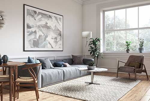 Moderne woonkamer met grijze hoekzetel, abstracte wanddecoratie, kamerplanten en een eethoek met houten meubels, verlicht door natuurlijk licht via een groot raam.