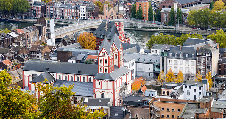 Aerial view of a European city with a red and white church featuring multiple spires, surrounded by buildings and autumn-colored trees, with a river and bridge in the background.