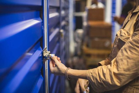 Personne verrouillant ou déverrouillant la porte d'une unité de stockage bleue avec un cadenas, avec d'autres unités et des boîtes empilées en arrière-plan.