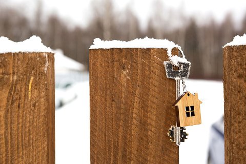 House keys with a wooden house keychain hanging on a snow-covered fence post, with a snowy landscape in the background