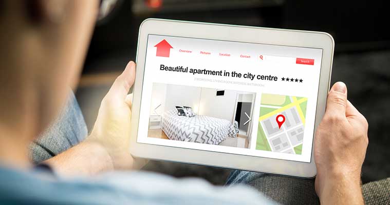Personne tenant une tablette affichant une page web d’un appartement en centre-ville, montrant une photo de chambre, des onglets de navigation et une carte avec une épingle rouge.